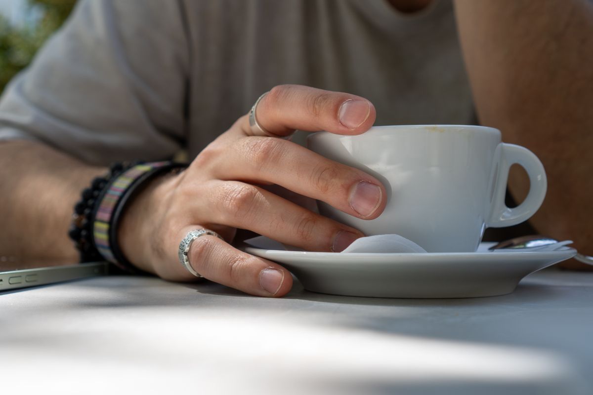 A hand with a natural manicure wearing two thin stackable gold bands, casually holding an iced coffee glass.